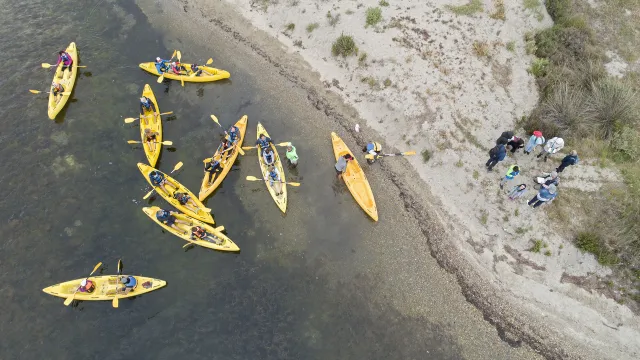 Vue aérienne de plusieurs kayaks jaunes sur une zone peu profonde, près d’une berge sableuse. Des participant·es embarquent ou débarquent tandis qu’un groupe observe depuis le rivage.